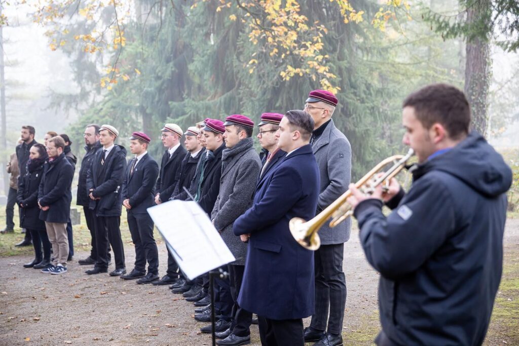 Timm Westermann und Henning Eggers und weitere Männer in Couleur und 2 Frauen beim Gedenken im Wald, eine Person spielt Trompete.