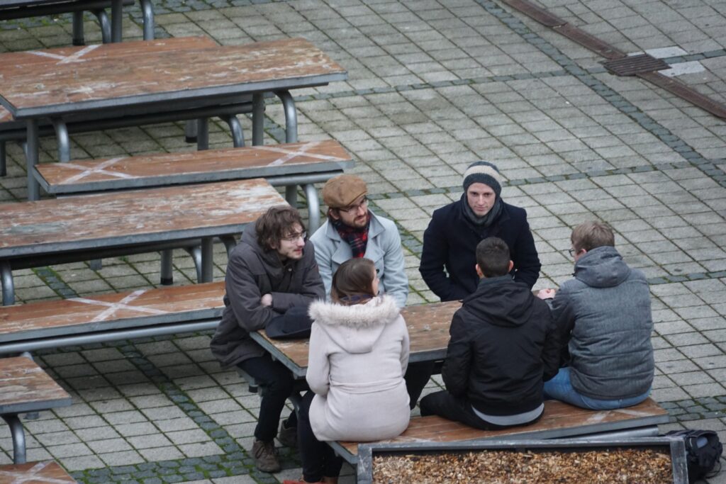 Sechs Personen sitzen draußen an einem Tisch auf dem Campus der Uni Jena und scheinen sich zu unterhalten.