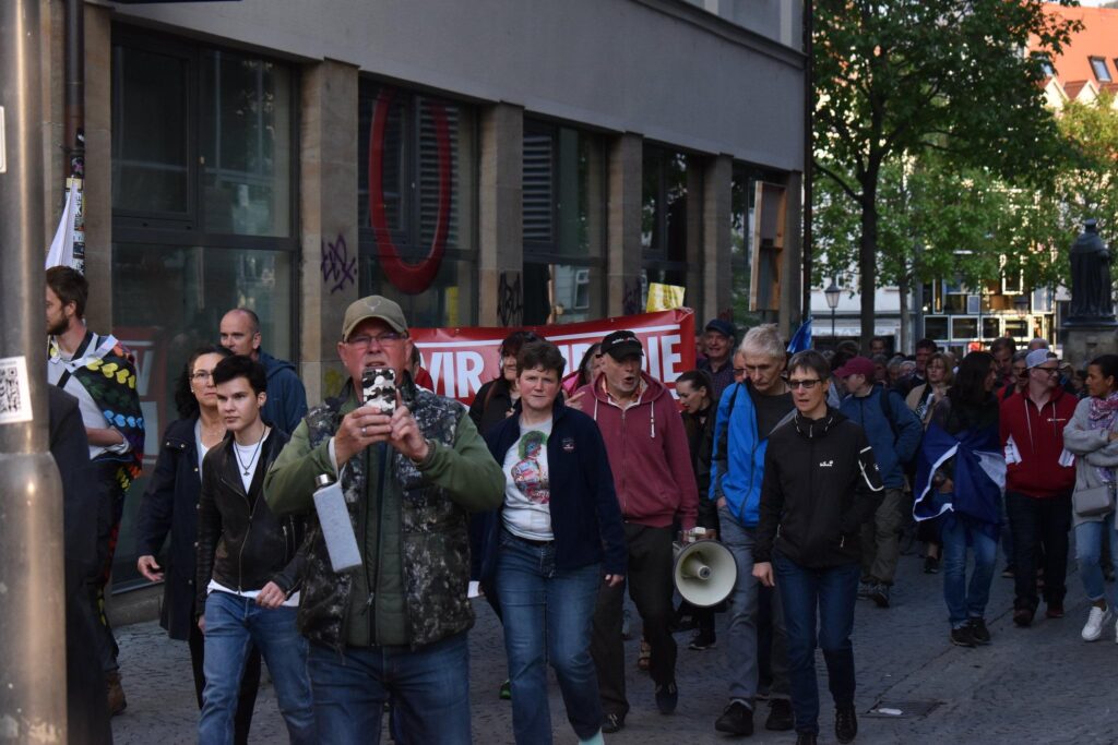 Bild von einem PandemieleugnerInnenaufmarsch in Jena. Bei dem Dante Riedel im vorderen Bereich zu sehen ist. Im Hintergrund ist ein Banner mit "Wir sind die rote Linie" zu sehen, wobei einige Buchstaben durch Personen verdeckt sind.