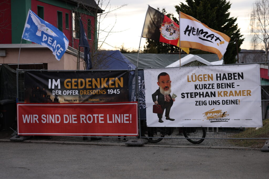 Foto von einem Zaun der als Sichtschutz vor dem Parteibüro aufgebaut ist. Daran sind Transparente und Fahnen befestigt. Auf den Transparenten steht: "In Gedenken der Opfer Dresdens 1945", "Wir sind die rote Linie" und "Lügen haben kurze Beine - Stephan Kramer zeig uns deine". Als Fahnen sind eine Peace Fahne mit einer weißen Taube, eine Fahne des Fürstentums Reuß, der Heimat und der Gerschen Jugend zu sehen.