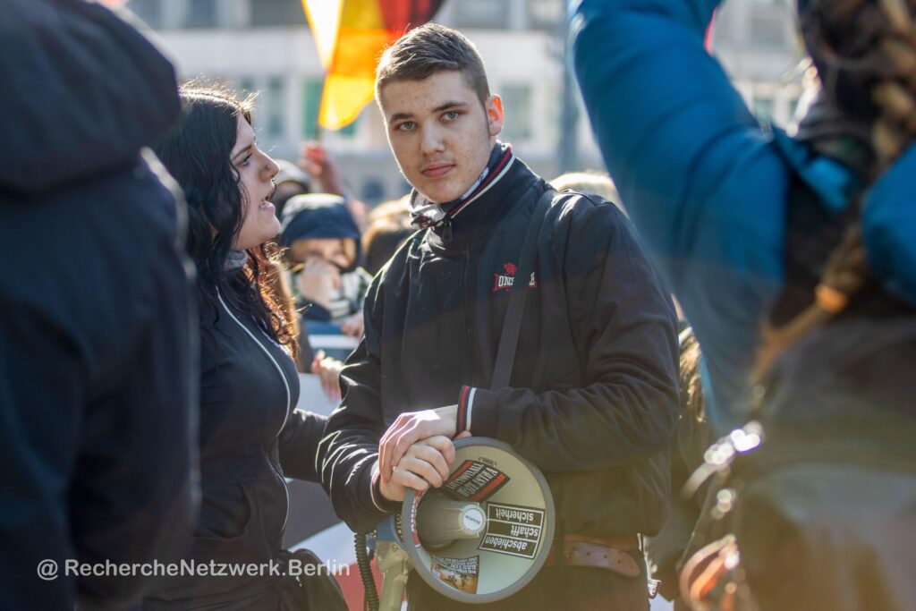 Foto auf dem Justin auf einem Neonaziaufmarsch in Berlin mit Megafon zu sehen ist. Das Megafon ist mit zahlreichen extrem rechten Stickern beklebt.