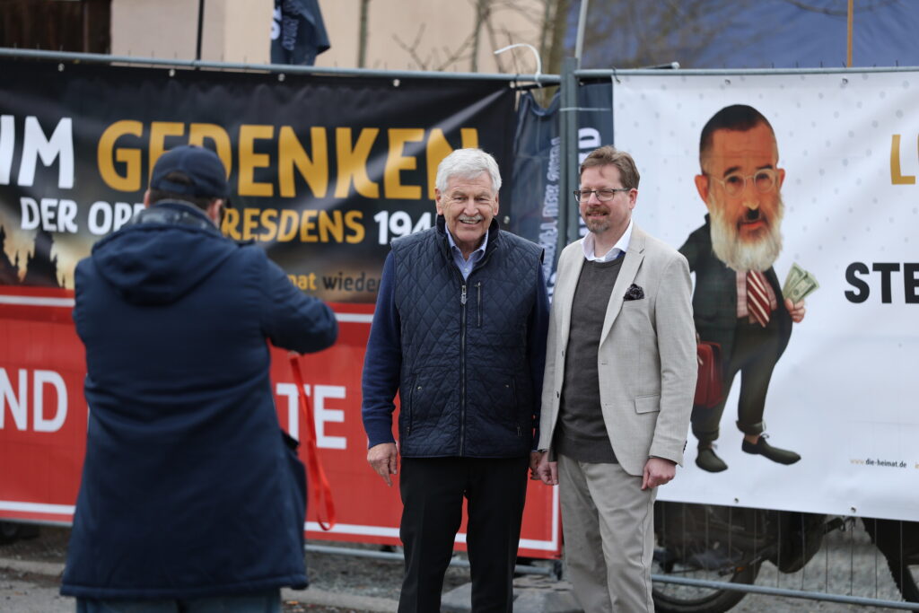 Udo Voigt und Peter Schreiber posieren vor dem Zaun vor dem neuen Parteibüro. Eine dritte Person macht ein Foto von ihnen.