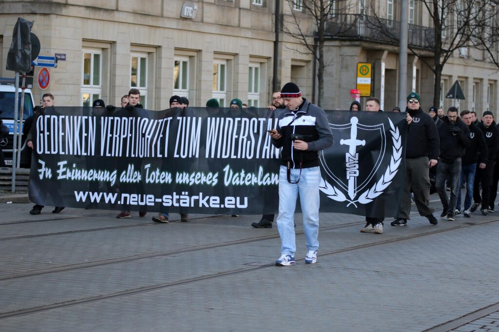Man sieht die Spitze eines Blocks im Neonaziaufmarsch in Dresden. Auf dem Fronttransparent steht "Gedenken verpflichtet zum Widerstand. Zur Erinnerung aller Toten unseres Vaterlands." Links am Transparent sieht man Etienne Klupp.