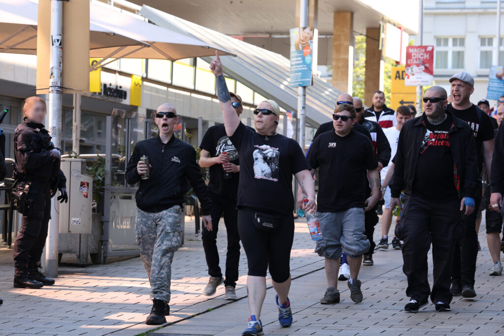 Man sieht eine Gruppe Neonazi-Skinheads mit Sonnenbrillen und schwarzer Kleidung in der Geraer Fußgängerzone. Mehrere Personen, darunter Pascal Chmielewski, grölen im Laufen.