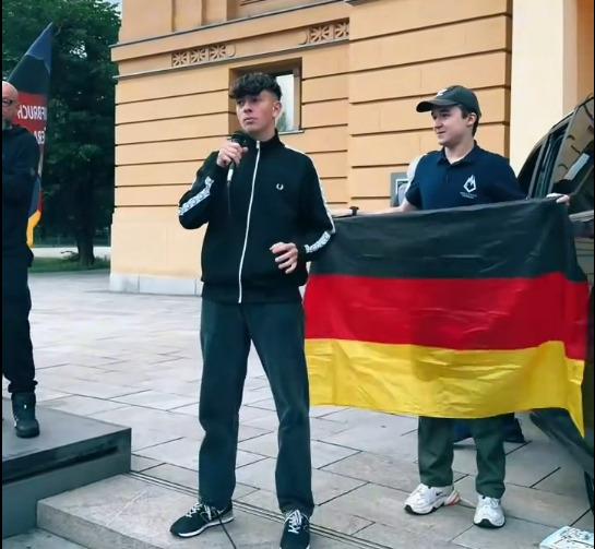 Man sieht Anthony Scholz in einer schwarzen Trainingsjacke am Mikrofon auf der Abschlusskundgebung der Geraer Montagsdemo vor dem Geraer Theater. Neben ihm hält ein junger Mann eine Deutschlandflagge.
