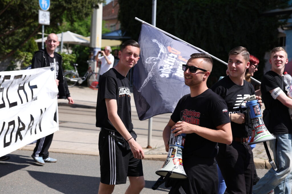 Vor einem Banner "Deutsche Jugend Voran" sieht man Anthony Scholz mit einem Shirt "Antifa-Antifa", Julian Milz mit einem Megafon und Justin Elbel mit einem Shirt "Nazibande".