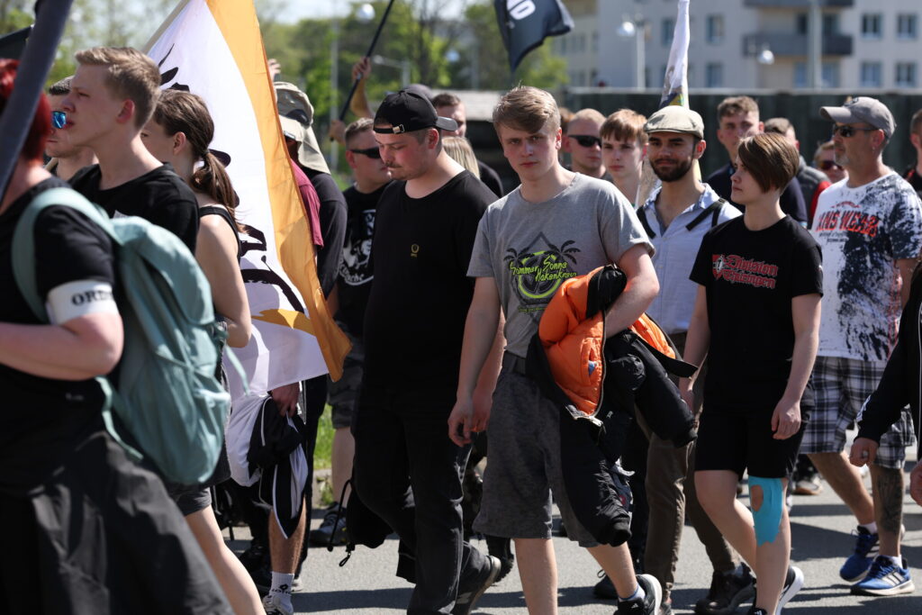 Man sieht mehrere junge Menschen im Naziaufmarsch von Gera am 1.5.2025. Hinter einer Flagge von "Die Heimat" läuft ein junger Mann mit einem grauen T-Shirt, auf dem ein rund stilisiertes Hakenkreuz abgebildet ist, über und unter dem steht: "Sommer, Sonne Hakenkreuz. Sieg fucking Heil." Dahinter läuft Caroline Hein mit einem schwarzen T-Shirt mit roter Aufschrift "Division Thüringen".