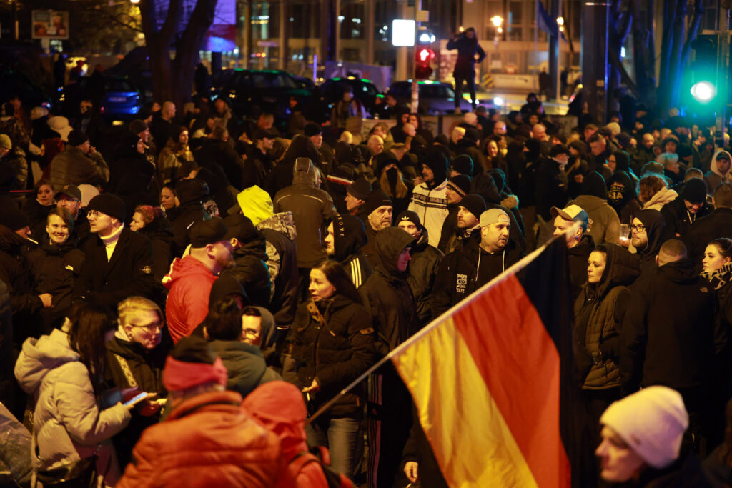 Man sieht Hunderte, oft junge Neonazis im Dunkeln auf dem Magdeburger Hasselbachplatz. Vorne wird eine Deutschlandfahne gehalten. Am linken Bildrand sieht man Caroline Hein lachend mit einer schwarzen Jacke.