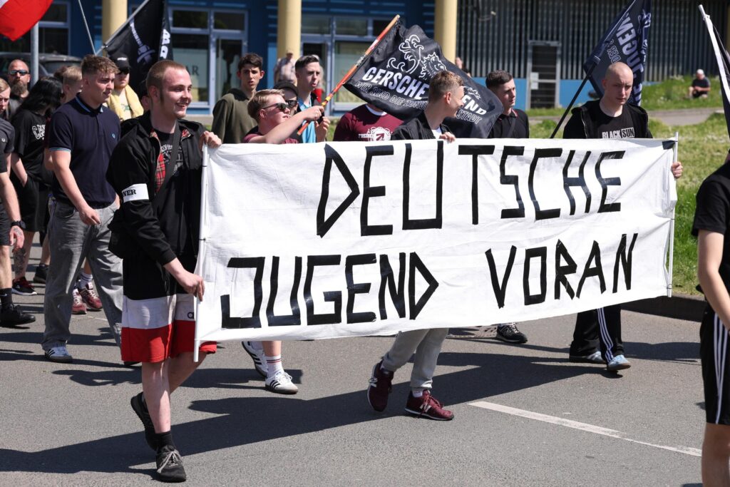 Foto des Neonaziaufmarsch in Gera am 1. Mai 2025. Drei Personen tragen ein Transparent mit "Deutsche Jugend voran", dahinter laufen Personen von denen zwei Personen Fahnen der "Gerschen Jugend" schwenken. Einer der Fahnenschwenker ist Vincent Thomas.