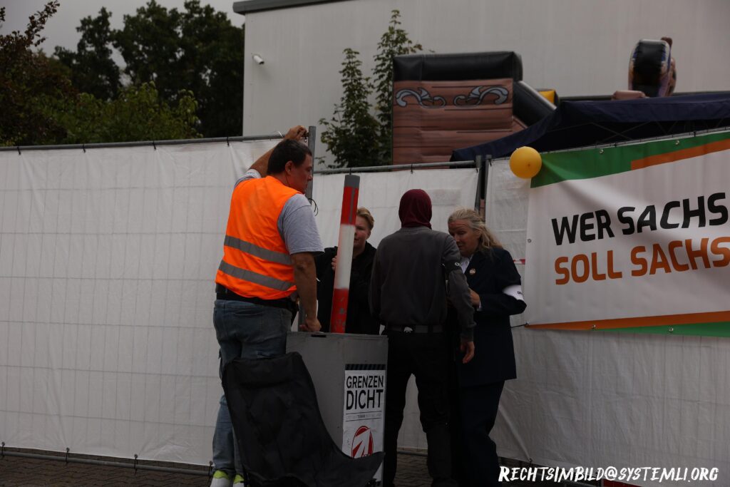 Foto vom Eingang der "Heimat"-Veranstaltung. Mehrere Bauzäune, die mit Planen bespannt sind, sind zur Abschirmung aufgestellt. An diesen hängt auch ein Transparent der "Freien Sachsen" und ein Luftballon. Hinter den Bauzäunen ist eine Hüpfburg zu sehen. Am Eingang steht neben drei weiteren Personen Vincent Thomas.