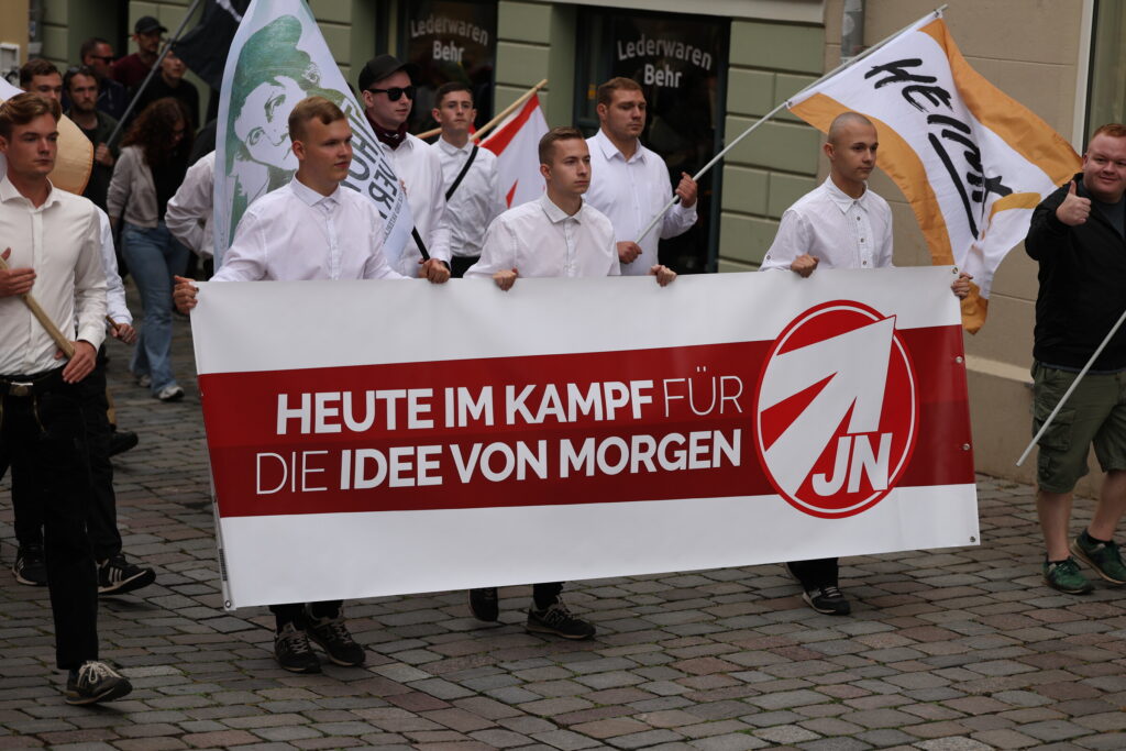 Foto des rassistischen Aufmarsch am 12.07.2025 in Apolda. Darauf sind mehrere junge Männer in weißen Hemden zu sehen. Diese tragen ein Transparent der JN mit dem Text "Heute im Kampf für die Idee von morgen" und Fahnen der "Heimat" und von "Thüringen verteidigen".