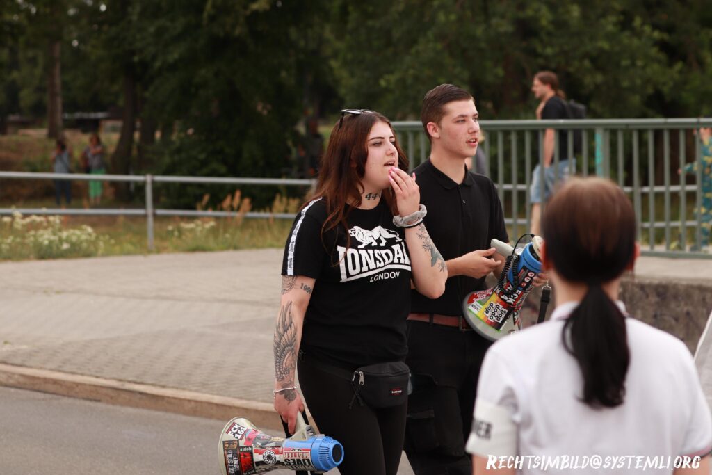 Foto eines queerfeindlichen Aufmarschs am 05.07.2025 in Falkensee. Darauf sind Stella Schmidt und Justin Eibel am Beginn des Aufmarschs zu sehen. Beide halten Megafone in der Hand, die mit zahlreichen rechten Stickern beklebt sind.