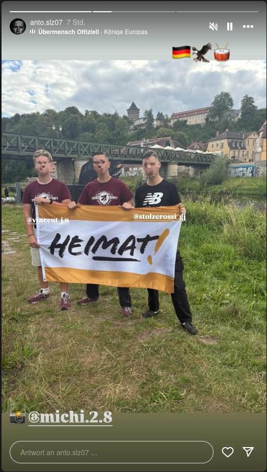 Instagram-Story des Accounts "anto.slz07". In dieser ist ein Foto von Vincent Thomas, Anthony Scholz und Justin Eibel zu sehen. Sie halten eine Fahne der "Heimat". Thomas und Scholz tragen T-Shirts der JN, Eibel trägt ein T-Shirt auf dem die Buchstaben "NB" im Stil von "New Balance" zu sehen sind. Unter dem Foto ist ein Fotoapparat Emoji und damit "@michi.2.8" zu sehen über den Fotos die Emojies: Deutschlandfahne, Adler und Trommel.
