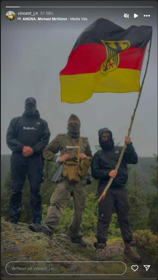 Instagram-Story des Accounts "vincent_j.n" mit einem Foto von Pablo Wolfer, Eric Vogelgesang und Vincent Thomas auf einem Felsen und im Hintergrund ein Wald. Alle drei sind mit Schlauchschals vermummt. Vogelgesang trägt eine Militäruniform, Thomas einen Schlauchschal der JN und eine schwarz-rot-goldene Fahne mit Bundesadler an einem Stock.