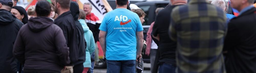 Foto von der Demonstration am 12.7.25 in Apolda. Es ist eine Person mit einem blauen AfD T-Shirt in der Mitte zu sehen. Im Hintergrund sieht man eine "Heimat"-Fahne, die geschwenkt wird.