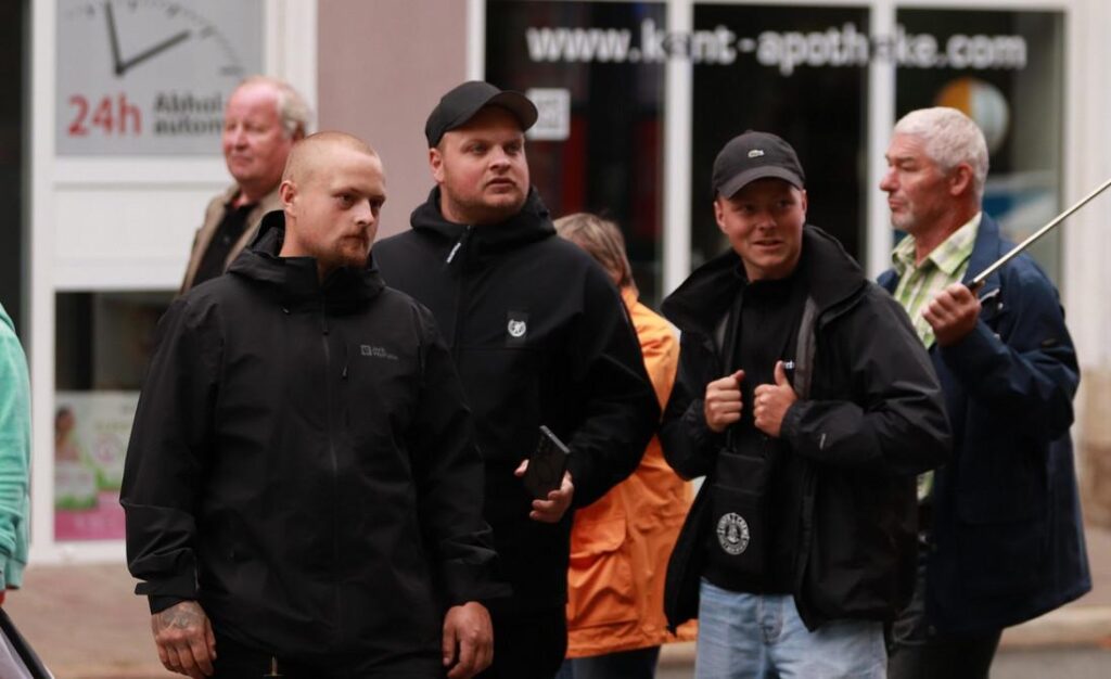 Foto von drei jugendlichen Neonazis in dunkler Kleidung bei der Demo am 12.7.25 in Apolda. Dahinter sind ältere Demoteilnehmende zu sehen, von denen einer eine Fahne trägt.