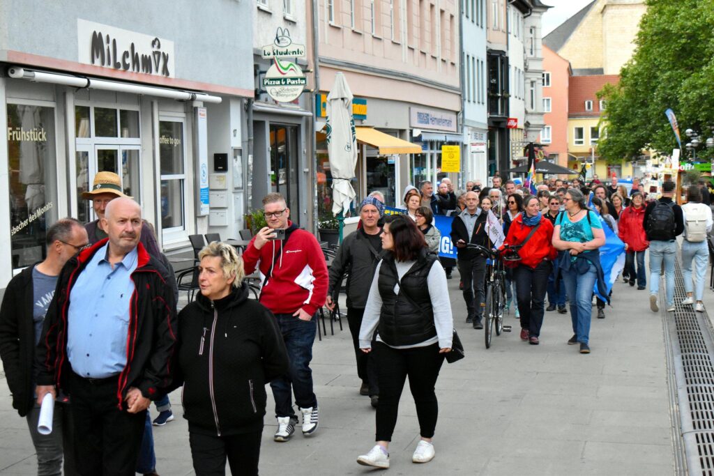 Foto eines PandemieleugnerInnenaufmarsch, der durch die Jenaer Johannisstraße läuft. Im Vordergrund läuft Volker Wagenhaus Hand in Hand mit einer anderen Person. Dahinter laufen einige weitere Personen, die ein Transparent tragen, dessen Beschriftung nicht lesbar ist.