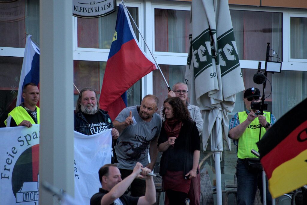 Foto von Corinna Gehre am 20.06.2022 in Gera. Gehre trägt rote und schwarze Kleidung und ist im gespräch mit Christian Klar. Um sie herum stehen andere Teilnehmende; auf der linken Seite der Neonazi Andy Fahl, auf der rechten in Presseweste und mit Streaming-Ausrüstung Andreas Thomä.