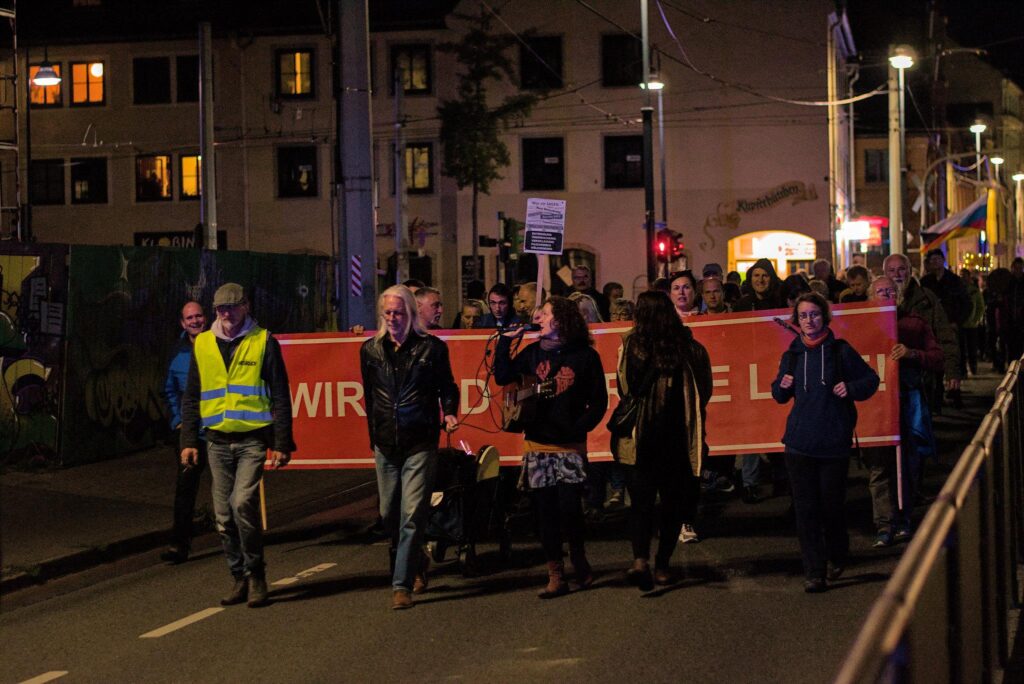 Foto von Corinna Gehre am roten Frontbanner eines PandemieleugnerInnen-Aufmarschs am 26.09.2022 in Jena. Sie wendet sich der Demo zu und spricht in ein Mikrofon, sie trägt zudem eine Gitarre umgehängt.