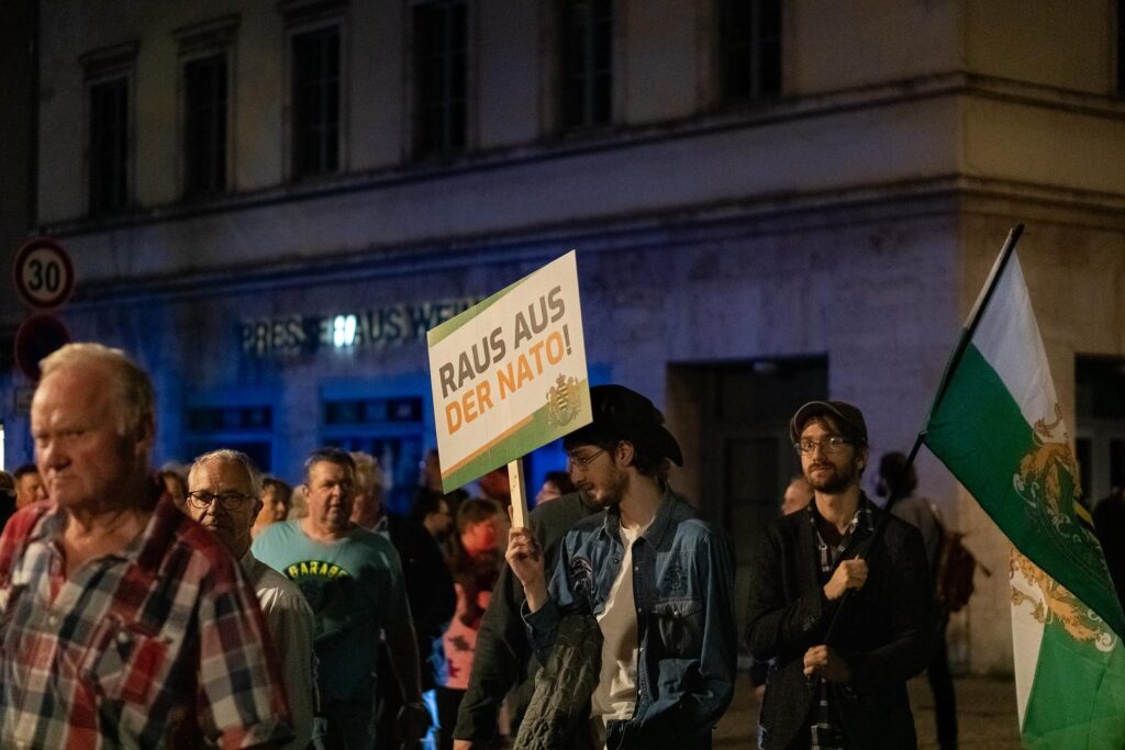 Foto von Michael Brauer bei einem verschwörungsideologischen Aufmarsch am 17.10.2022 in Weimar. Brauer steht mit schwarzer Jacke und Kappe am rechten Bildrand. Er hat eine Fahne vom ehemaligen Königreich Sachsen in den Händen. Im Vordergrund ist ein anderer Teilnehmer mit einem Schild zu sehen, auf dem „Raus aus der Nato!“ steht.