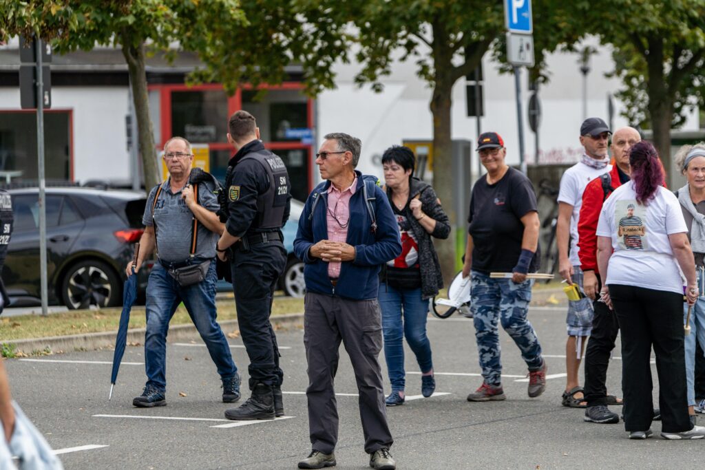 Foto von Angela Lehmann bei dem vom Neonazi und „Heimat“-Mitglied Christan Klar organisierten extrem rechten Aufmarsch am 3. Oktober 2023. Lehmann trägt ein schwarzes Shirt, eine Hose in Tarnfarben und eine Kappe. Sie trägt ihre Trommel in der rechten und ihre Stöcke in der rechten Hand. Um sie herum unterhalten sich andere Teilnehmende des Aufmarsches, links von ihr steht ein Polizist mit dem Rücken zur Kamera.