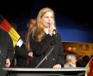 Foto von Wiebke Muhsal an einem Redner*innenpult. Sie hält ein Mikrofon in der Hand. Im Hintergrund steht eine Person mit einer schwarz-rot-goldenen Fahne.