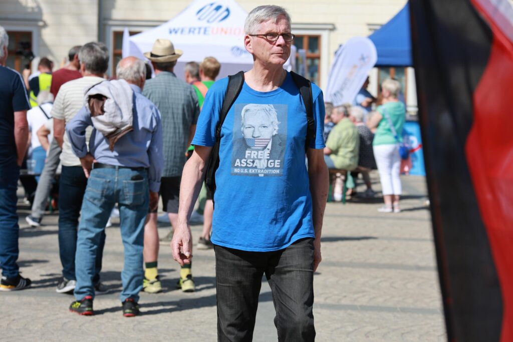 Foto von Winfried Merkel am 1. Mai 2024 auf Kundgebung von „Freies Thüringen“ und „Heimat“ in Sondershausen. Er trägt ein blaues T-Shirt mit einem Bild von Julian Assange mit der Auschrift „free Julian Assange“. Im Hintergrund sind andere Teilnehmende der Kundgebung zu sehen. Am rechten Bildrand ist eine Deutschlandfahne.
