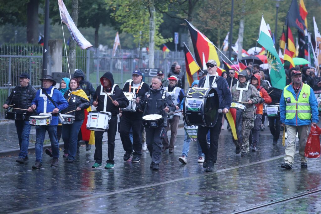 Foto von Angela Lehmann bei dem vom Neonazi und „Heimat“-Mitglied Christan Klar organisierten extrem rechten Aufmarsch am 3. Oktober 2024. Angela Lehmann ist auf der rechten Bildhälfte zusehen. Sie spielt eine Trommel, an der eine Deutschlandfagge hängt.Es regnet und die DemonstrantInnen tragen Regenkleidung, einige tragen Deutschland- und Sachsenfahnen mit sich.