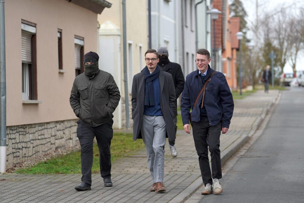 Foto von Friedrich Hunger und Timm-Albert Westermann bei den Studientagen in Schnellroda am 25.01.2025. Hunger läuft in der Mitte und trägt Brille, ein blaues Oberteil, eine graue Hose und einen grauen Mantel. Rechts von ihm ist Westermann zu sehen, hinter und links von Hunger laufen zwei vermummte, dunkel gekleidete Personen.
