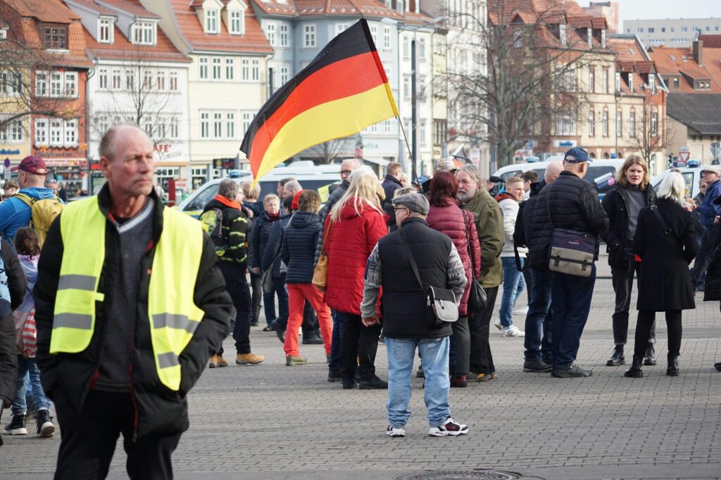 Foto von Uwe Schroth beim AfD-Wahkampfabschluss am 22.02.2025 in Erfurt. Im Hintegrund ist die Erfurter Altstadt zu sehen, davor stehen zahlreiche Erwachsene. In der Bildmitte weht eine Deutschlandfahne. Links im Vordergrund des Bildes steht Uwe Schroth in einer schwarzen Jacke und einer neongelben Ordnerweste.