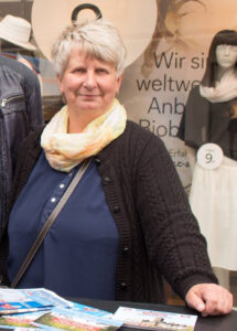 Foto von Birgit Schild auf dem diese an einem Tisch steht auf dem AfD-Flyer liegen. Im Hintergrund ist das Schaufenster eines Kleidungsgeschäfts zu sehen.