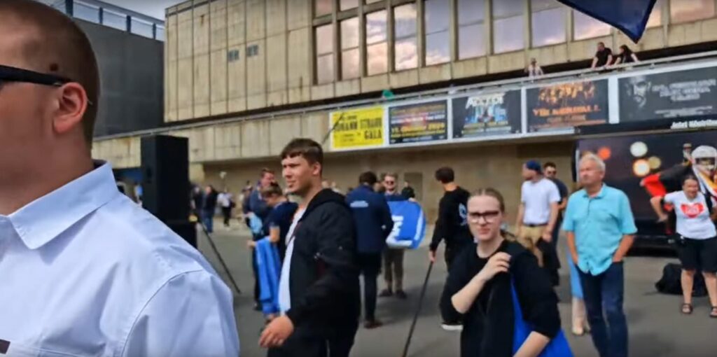 Screenshot von einem Livestream, auf dem Julian und Haily-Marie Kitz am 24.06.2024 in Gera zu sehen sind. Julian trägt ein weißes T-Shirt und eine schwarze Jacke, Kitz eine blaue Tasche, einen schwarzen Pullover und eine große blaue Tasche.