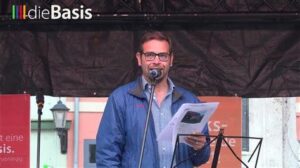 Foto von Karsten Geschwandter auf einer Bühne mit Mikrofon. In der linken oberen Ecke des Fotos ist das Wasserzeichen "dieBasis" zu sehen.