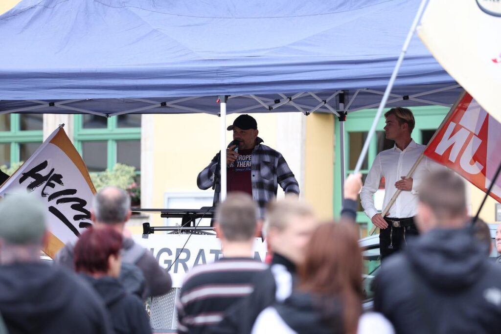 Foto von Christian Klar und Etienne Klupp am 12.7.25 in Apolda. Beide stehen unter einem Pavilon. Klar hält ein Mikrofon in der Hand, in das er spricht. Klupp steht daneben und trägt eine Fahne der JN.
