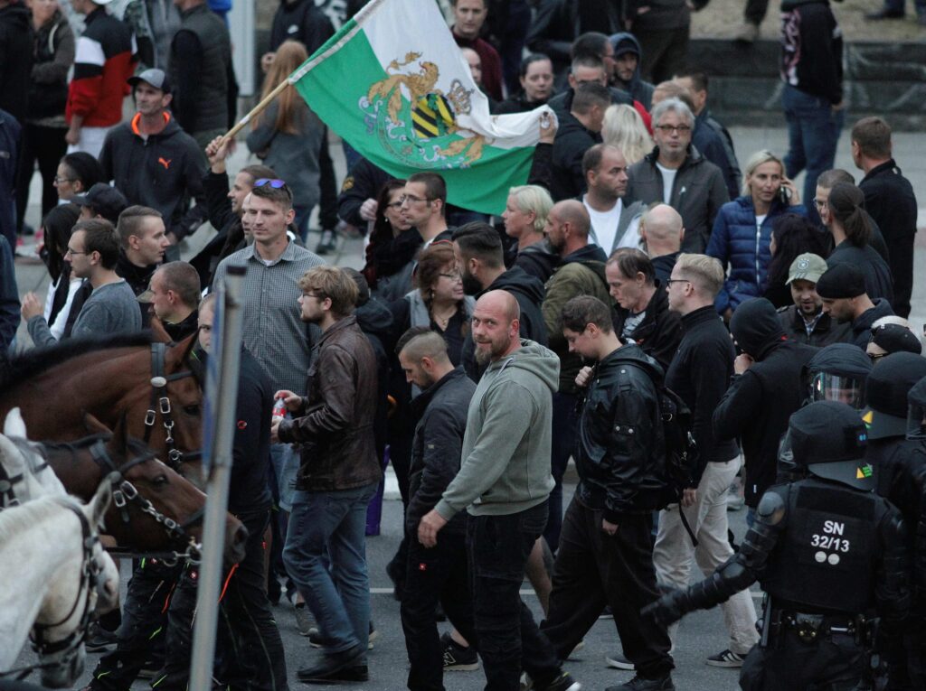 Wasielewski und Marcel Waschek in Chemnitz am 1.9.2018. Wasielewski ist am rechten Bildrand zu sehen und trägt Brille sowie einen grauen Pullover. Die beiden sind in einem Pulk anderer Teilnemender in dunkler Kleidung unterwegs. Eine Person schwenkt eine Sachsenfahne. Am linken Rand sind zudem die Köpfe von vier Pferden zu sehen.