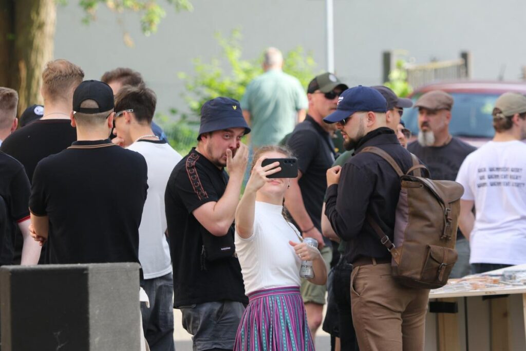 Kundgebung der Identitären. Mittig steht Robert Brandt in schwarzem Shirt und mit dunkelblauem Fischerhut, die Hand halb das Gesicht verdeckend. Rechts davon macht seine Freundin Freya Schöps, langer Rock und Flechtzöpfe, ein Selfie. Rechts der beiden steht Leonardo Lekhi mit sonnenbrille und dunkelblauer Cap.