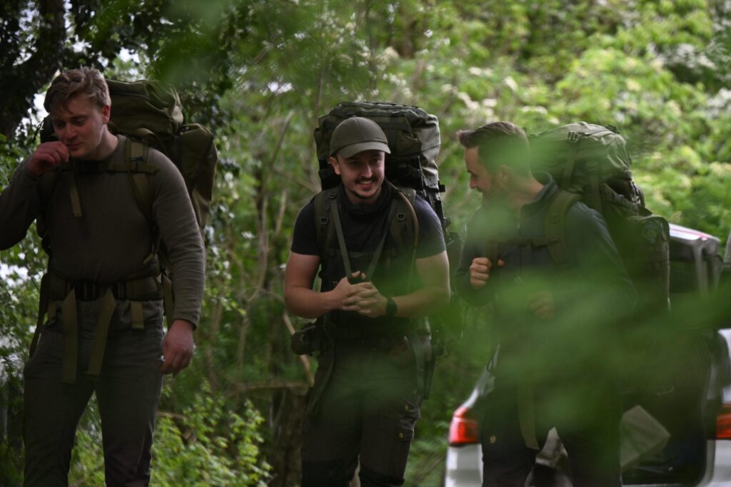 Drei junge Männer wandern nebeneinander durch einen Wald. Mittig sieht man Niklas Aust in dunkelgrüner Tarnkleidung, mit Camouflagerucksack und -mütze lachend.