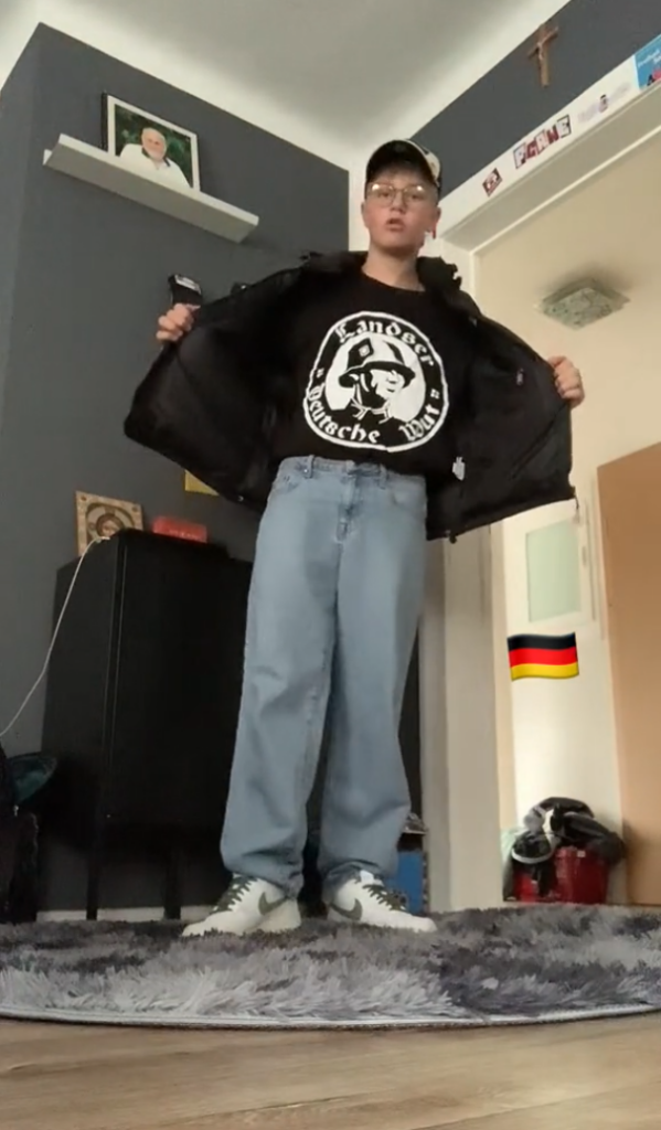 Screenshot aus einem Video von Pablo Kühn: Er steht in seinem Zimmer mit heller Cap und einem schwarzen „Landser“-Shirt mit Stahlhelm-Symbol. 