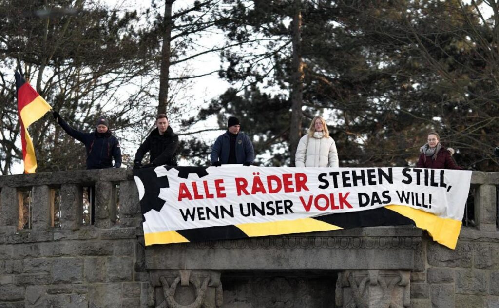 An der Empore am Erfurt Stadtpark stehen mehrere junge Leute hinter einem entrollten Banner mit der Aufschrift „Alle Räder stehen still, wenn unser Volk das will!“. Die Person ganz links hält eine Deutschlandfahne