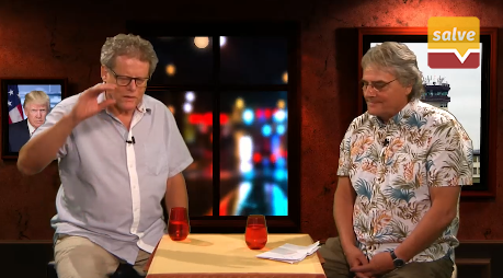 Man sieht Norbert Dotterweich und Klaus-Dieter Böhm-Schneider in kurzärmligen Hemden im Salve TV Studio im Gespräch. Die beiden stehen sich an einem kleinen Holztisch gegenüber, auf diesem sind zwei organgene Gläser sowie ein Stapel Papiere platziert, die Böhm-Schneider vor sich abgelegt hat. Er trägt ein buntes Hawaihemd und eine schwarze Uhr am linken Handgelenk, sein Blick wandert nach unten zu seinen Notizzetteln. Dotterweich ihm gegenüber trägt ein weites hellblaues Hemd, sein rechter Arm ist im Redeschwall leicht erhoben. Links hinter ihm ist auf einem Bildschirm oder Bild Donald Trump vor einer US-Fahne zu erkennen.