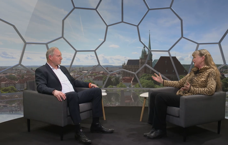 Wolfgang Lauerwald und Marion Schneider sitzen im kuppelförmigen Studio von Salve TV. Im Hintergrund sieht man die Dächer der Erfurter Innenstadt und den Dom. Schneider hat die Hände erhoben, während sie spricht.
