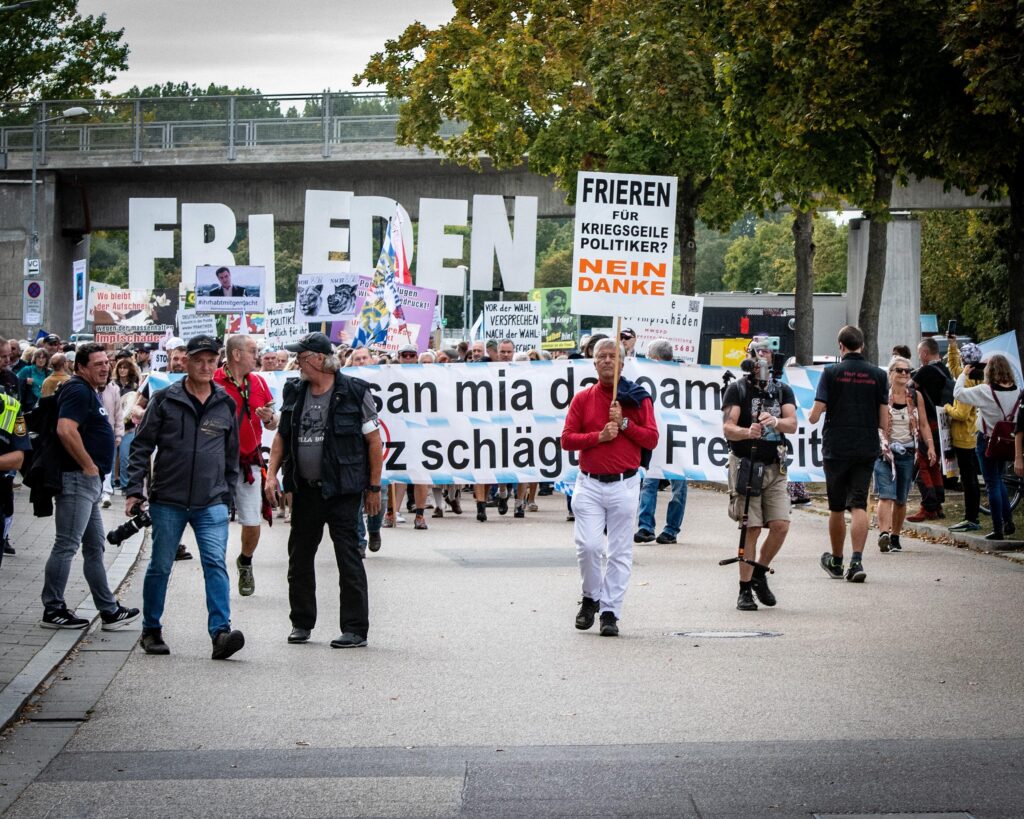 Man sieht die Front einer Demonstration, in der Mitte des Bildes läuft Pfaff mit rotem Pullover und weißer Hose. Er hält ein Schild mit folgender Aufschrift: Frieren für kriegsgeile Politiker? Nein danke. Im Hintergrund ist das Fronttransparent zu sehen. Dahinter halten Teilnehmende die Buchstaben des Wortes 'Freiden' in die Höhe.