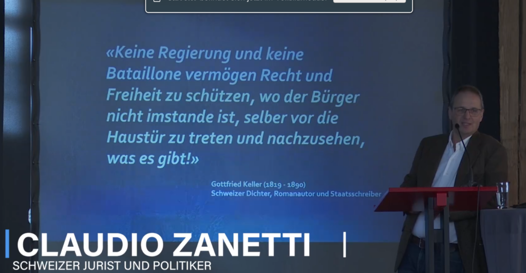 Ein Screenshot von Zanetti beim Kongress 2025. Am rechten Rand des Bildes lehnt Zanetti am Rednerpult und blickt ins Publikum. Hinter ihm sieht man eine Folie seiner Präsentation mit folgender Aufschrift: Keine Regierung und keine Bataillone vermögen Recht und Freiheit zu schützen, wo der Bürger nicht imstande ist, selber vor die Haustür zu treten und nachzusehen, was es gibt! Gottfried Keller (1819-1890). Schweizer Dichter, Romanautor und Staatsschreiber. Am unteren Bildrand ist eingeblendet: "Claudio Zanetti, Schweizer Jurist und Politiker"