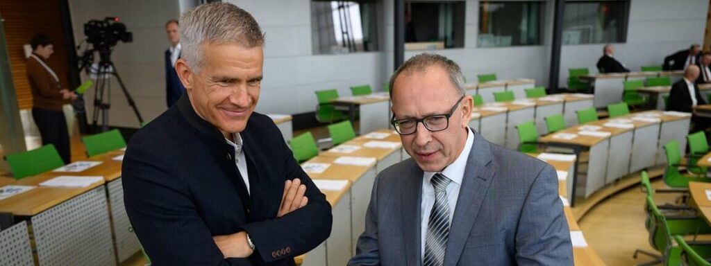 Berger und Urban stehen im sächsischen Landtag und unterhalten sich. Berger steht links, trägt eine dunkle Jacke und lächelt. Urban steht rechts und trägt einen grauen Anzug und Brille. Im Hintergrund sieht man die grünen Stühle des Landtages sowie eine laufende Kamera.