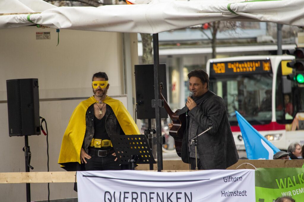 Krengel steht auf einer Bühne mit weißem Dach und hält eine Gitarre sowie ein Mikrofon, in das er spricht. Neben ihm steht ein Mann in gelber Verkleidung. An der Bühne hängt ein Transparent mit der Aufschrift: "Querdenken".
