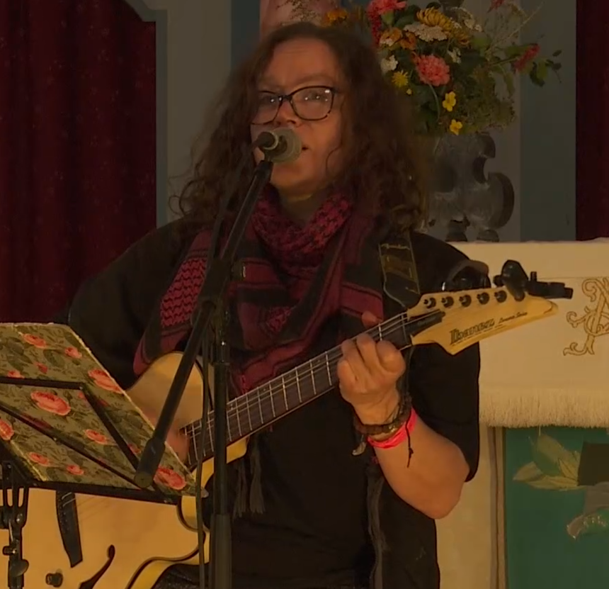 Gehre spielt Gitarre und singt. Vor sich hat sie einen Notenständer mit geblümter Mappe. Sie trägt ein schwarzes Kleid und eine rote Kufiyah. Im Hintergrund sieht man einen roten Vorhang und einen Blumenstrauß.