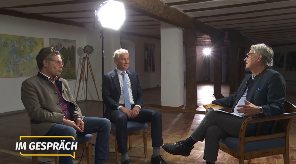 Dehm sitzt auf der linken Seite, in der Mitte des Bildes ist Höcke, rechts von ihnen Böhm, der gerade spricht und die beiden Gäste ansieht. Sie drei sitzen in einem großen Raum, der im Fachwerk-Stil gebaut ist. Sie werden von einer großen Studiolampe ausgeleuchtet. In der linken unteren Ecke steht: "Im Gespräch".