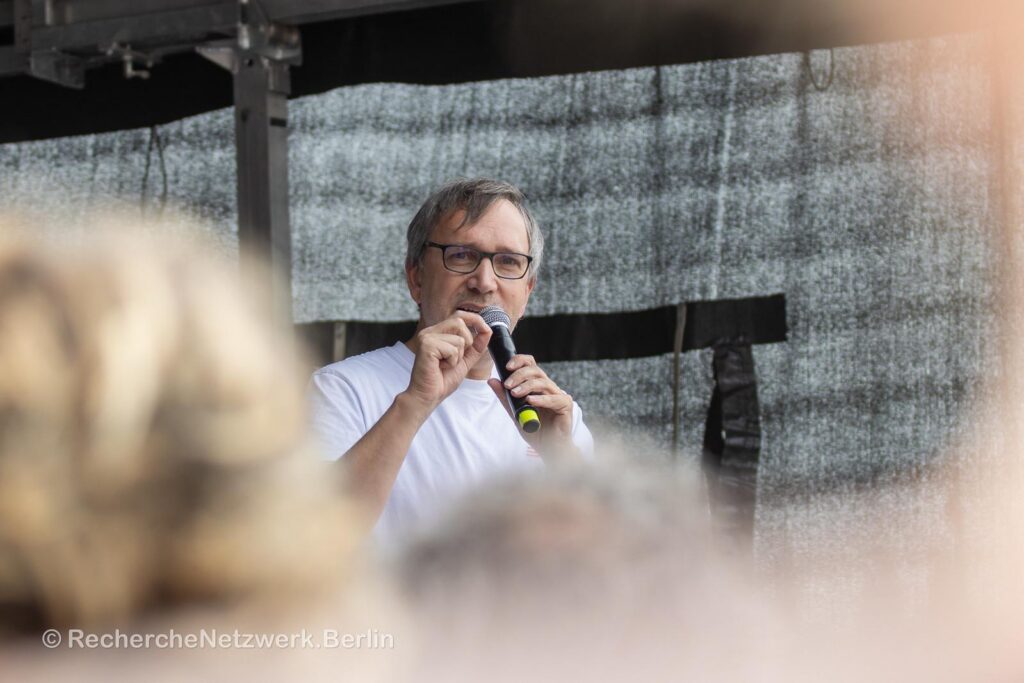 Ralf Ludwig trägt ein weißes Shirt und steht vor einem schwarz verhangenen Hintergrund. Er spricht in ein Mikrofon. Im Vordergrund sind verschwommen die Köpfe der Menschen zu sehen, die ihm zuhören.