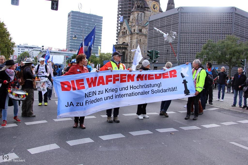 Christiane Reymann hält mit drei anderen Personen ein Transparent. Auf diesem steht: "Die Waffen nieder. Nein zu Krieg und Hochrüstung! Ja zu Frieden und internationaler Solidarität!." Hinter Reymann stehen noch weitere TeilnehmerInnen. Auf der linken Seite sind Leute mit Trommeln zu sehen, welche in Deutschlandfarben behangen sind.