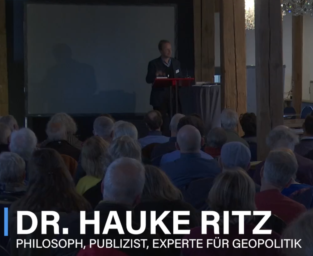 EIn Screenshot von Ritz auf dem Kongress 2025. Er steht in einem grauen Anzug am Rednerpult und blickts ins Publikum. Im Vordegrund sieht man die Hinterköpfe der Zuhörenden, die meisten haben graue Haare. Unten im Bild steht: "Dr. Hauke Ritz. Philosoph, Publizist, Experte für Geopolitik".
